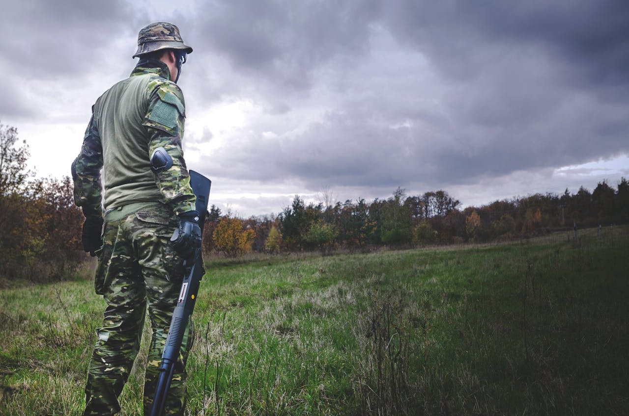 A soldier in camouflage with a rifle stands in a grassy field under a cloudy sky.